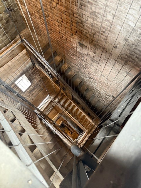 A view looking down into an industrial stairwell with brick walls, metal railings, and wooden steps.