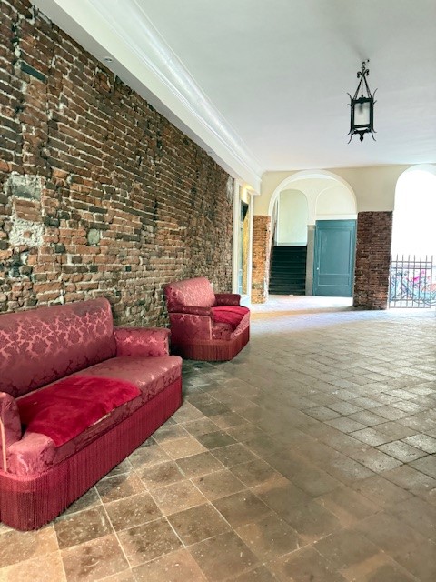 Two red upholstered sofas in a hallway with exposed brick walls and a tiled floor, featuring an overhead lantern and a doorway leading to a staircase.