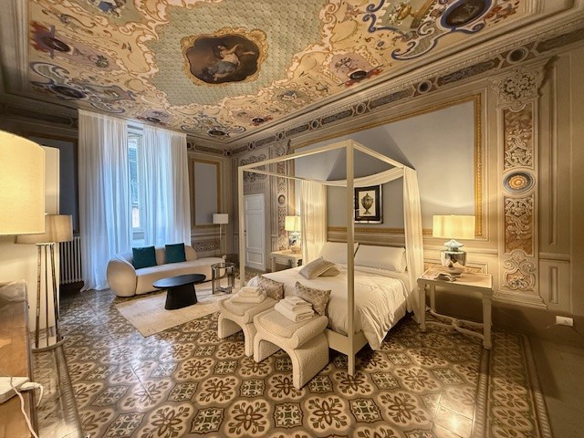 Elegant bedroom featuring a four-poster bed, large windows with sheer curtains, and ornate wall decorations. The room includes modern furniture like a circular sofa and a coffee table, with patterned tile flooring adding to the decor.