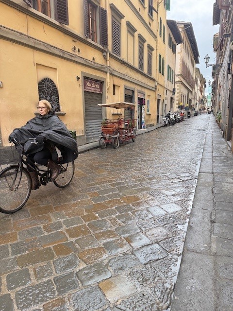 A cyclist wearing a poncho rides down a cobblestone street lined with buildings and parked bicycles.