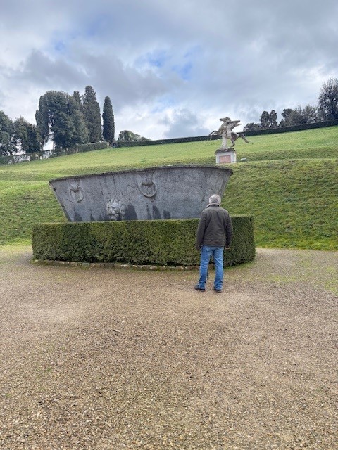 A person stands in front of a large stone basin surrounded by a circular hedge in a garden, with trees and a statue visible in the background under a cloudy sky.