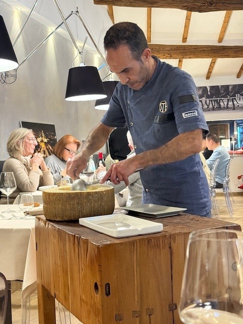 A chef skillfully preparing a dish at a wooden table in a cozy restaurant setting, with diners seated in the background.