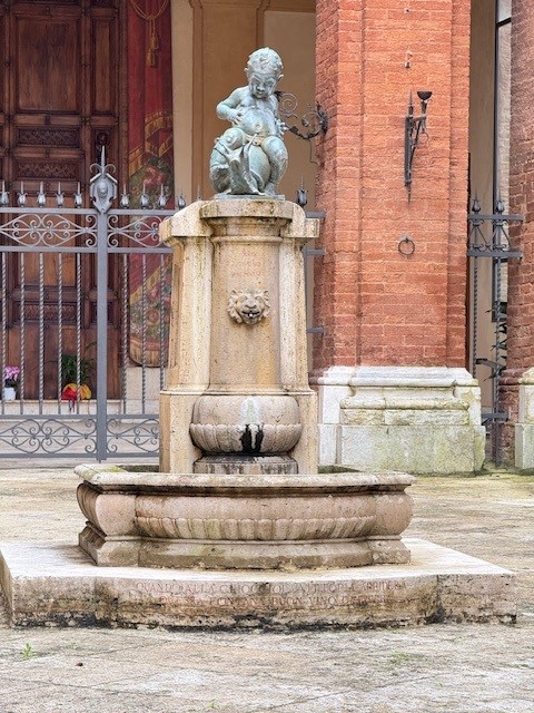 A stone fountain featuring a bronze statue of a cherub atop a decorative pedestal, set against a backdrop of an ornate gate and brick wall.