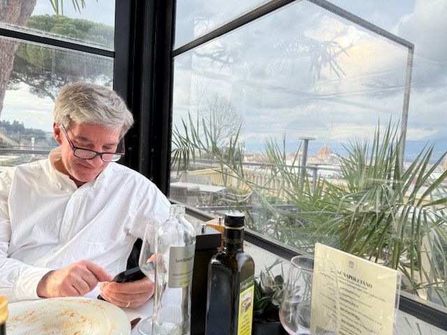 A man in a white shirt sitting at a table looking at his phone, with a view of a city landscape through large glass windows, surrounded by indoor plants.